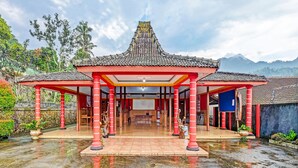 Un edificio tradicional con tejado de tejas y pilares rojos, rodeado de vegetación y un paisaje montañoso.