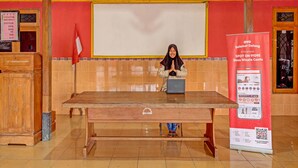 Una persona sentada a una mesa de madera con una computadora portátil, en una habitación con paredes rojas, una pizarra grande y retratos enmarcados en la pared.