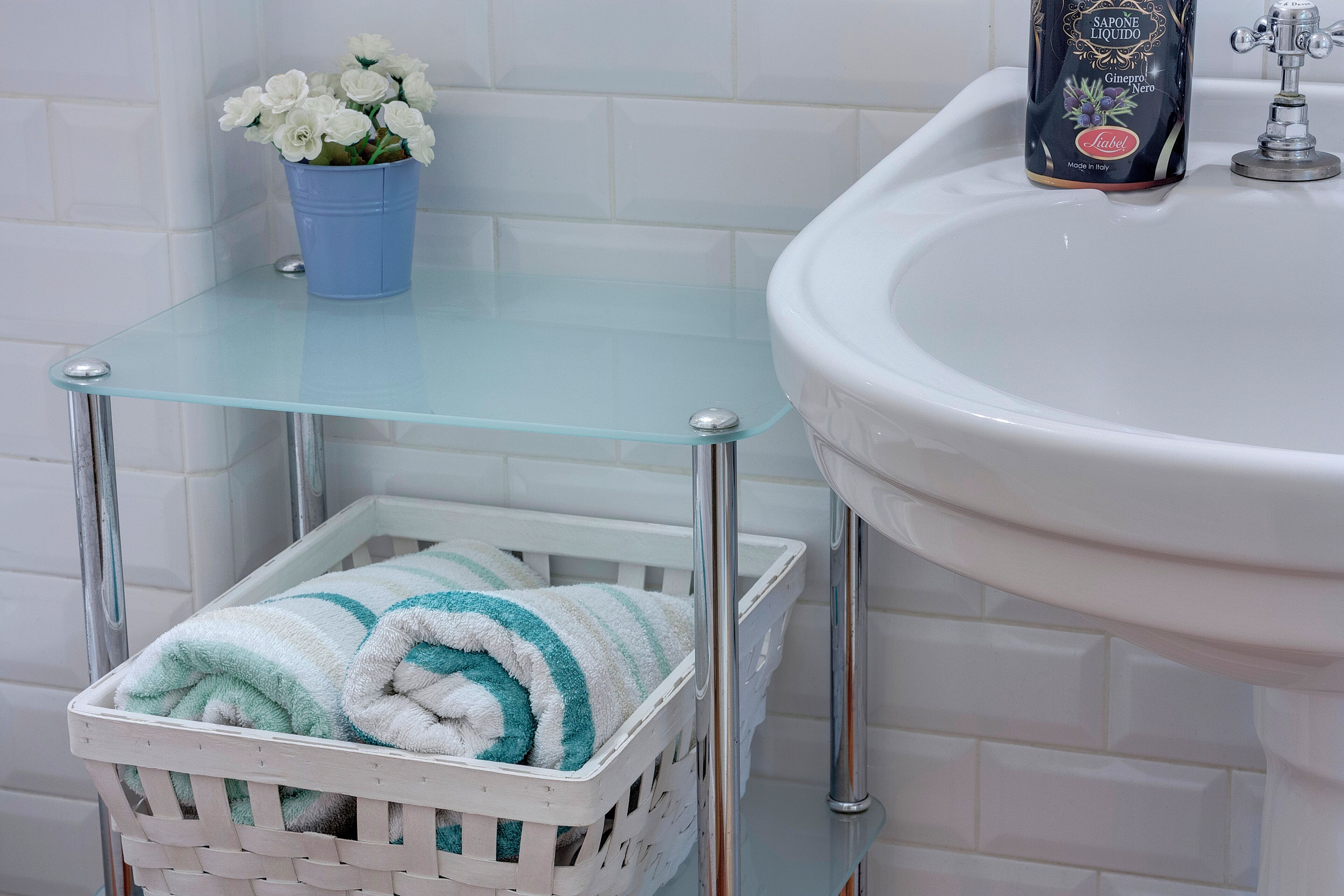 A bathroom with a glass shelf holding a blue bucket with white flowers, a white basket with a striped towel, and a white sink with a soap dispenser.