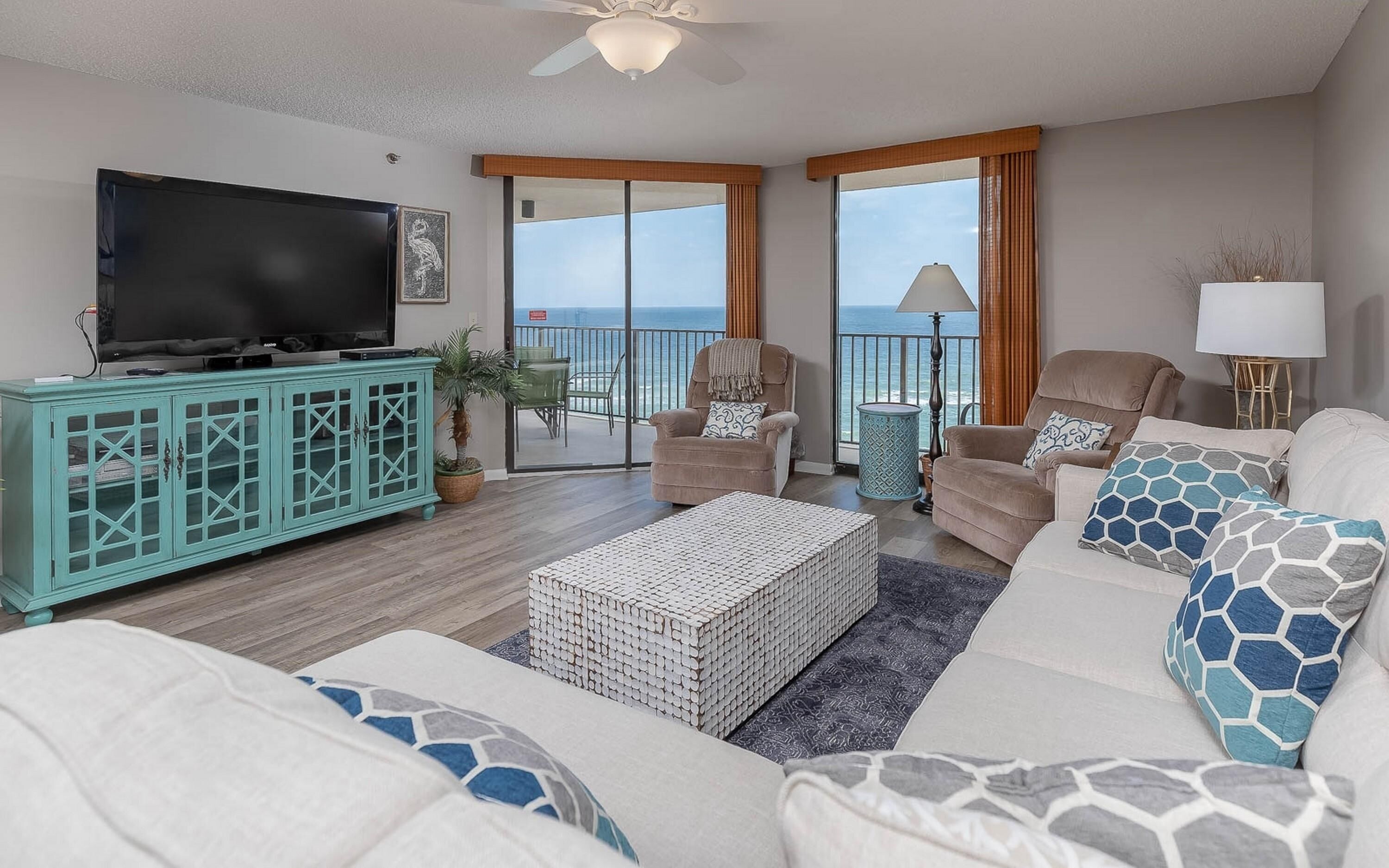 A spacious living room with a large flat-screen TV, teal-colored cabinet, two armchairs, a beige sofa with patterned cushions, and a view of the ocean through large sliding doors.