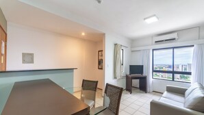 Een moderne woonkamer met een glazen eettafel, twee rieten fauteuils, een bank, een televisie op een dressoir en een groot raam met uitzicht op de stad.