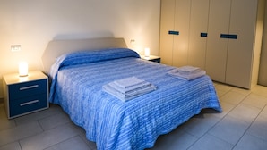 A neatly made bed with a blue striped bedspread and two folded towels on the side. There is a nightstand with a lamp and a drawer. A large wardrobe is visible in the background. - Brand New 2-bed Apartment With Patio (Endine Gaiano)