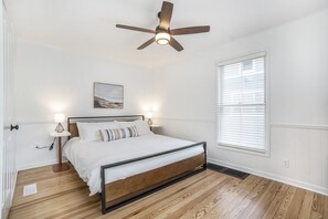 Ein Schlafzimmer mit Holzboden, einem Bett mit weißer Bettwäsche, einem Deckenventilator, Nachttischlampen und einem Fenster mit Jalousien.