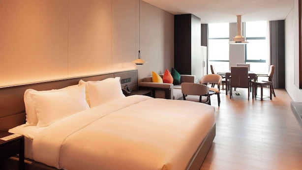 Une chambre d’hôtel moderne dotée d’un grand lit, d’un canapé, d’une table à manger avec des chaises et offrant une vue sur la ville grâce à de grandes fenêtres.