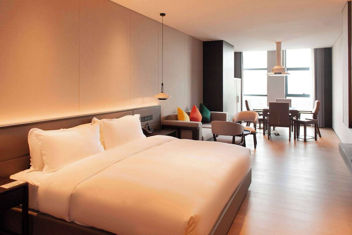 Ein modernes Hotelzimmer mit einem großen Bett, einer Couch, einem Esstisch mit Stühlen und Blick auf die Stadt durch große Fenster.