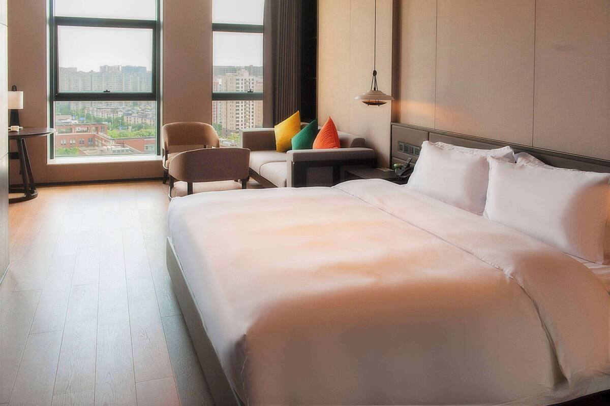 Ein Hotelzimmer mit einem großen Bett, einer Couch, einem Sessel und einem Schreibtisch. Große Fenster bieten einen Blick auf die Gebäude gegenüber.