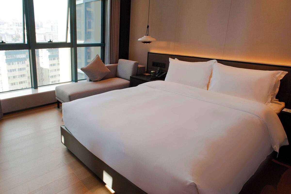 Ein Hotelzimmer mit einem großen Bett, einem Nachttisch, einem Sessel und einem Fenster mit Blick auf die Stadt.