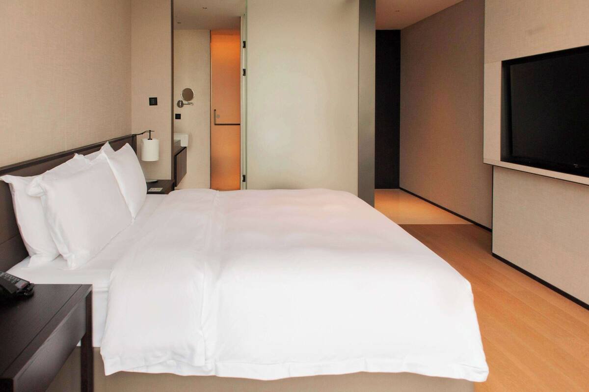 Ein Hotelzimmer mit einem großen Bett, einem an der Wand befestigten Fernseher, einem Nachttisch mit Lampe und einer Tür zu einem weiteren Raum.