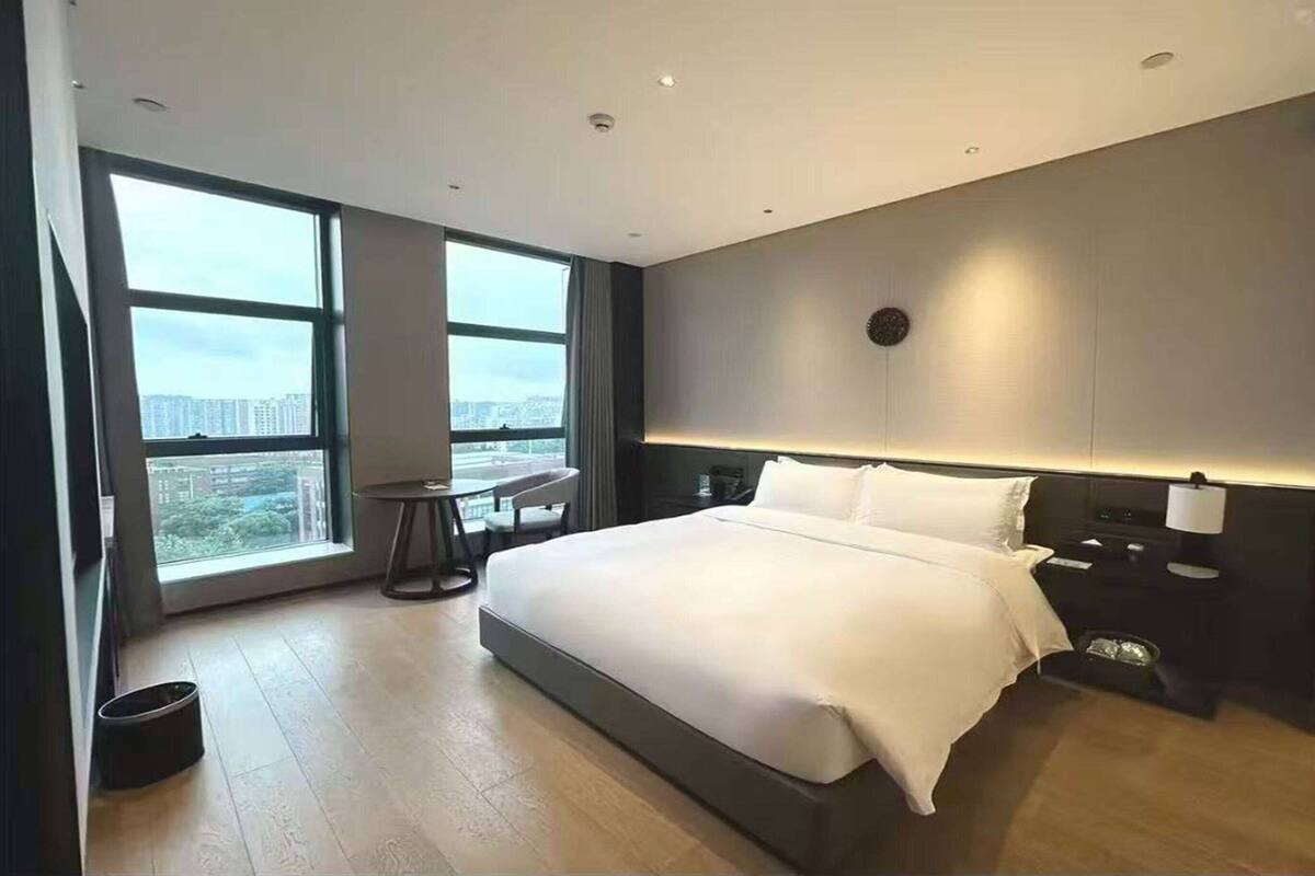 Ein modernes Hotelzimmer mit einem großen Bett, einem kleinen Tisch und Blick auf die Stadt durch große Fenster.