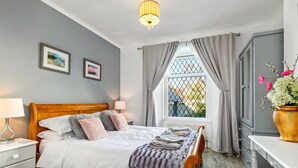 A bedroom with a wooden bed, a bedside table, a lamp, a window with curtains, and a view of greenery outside.