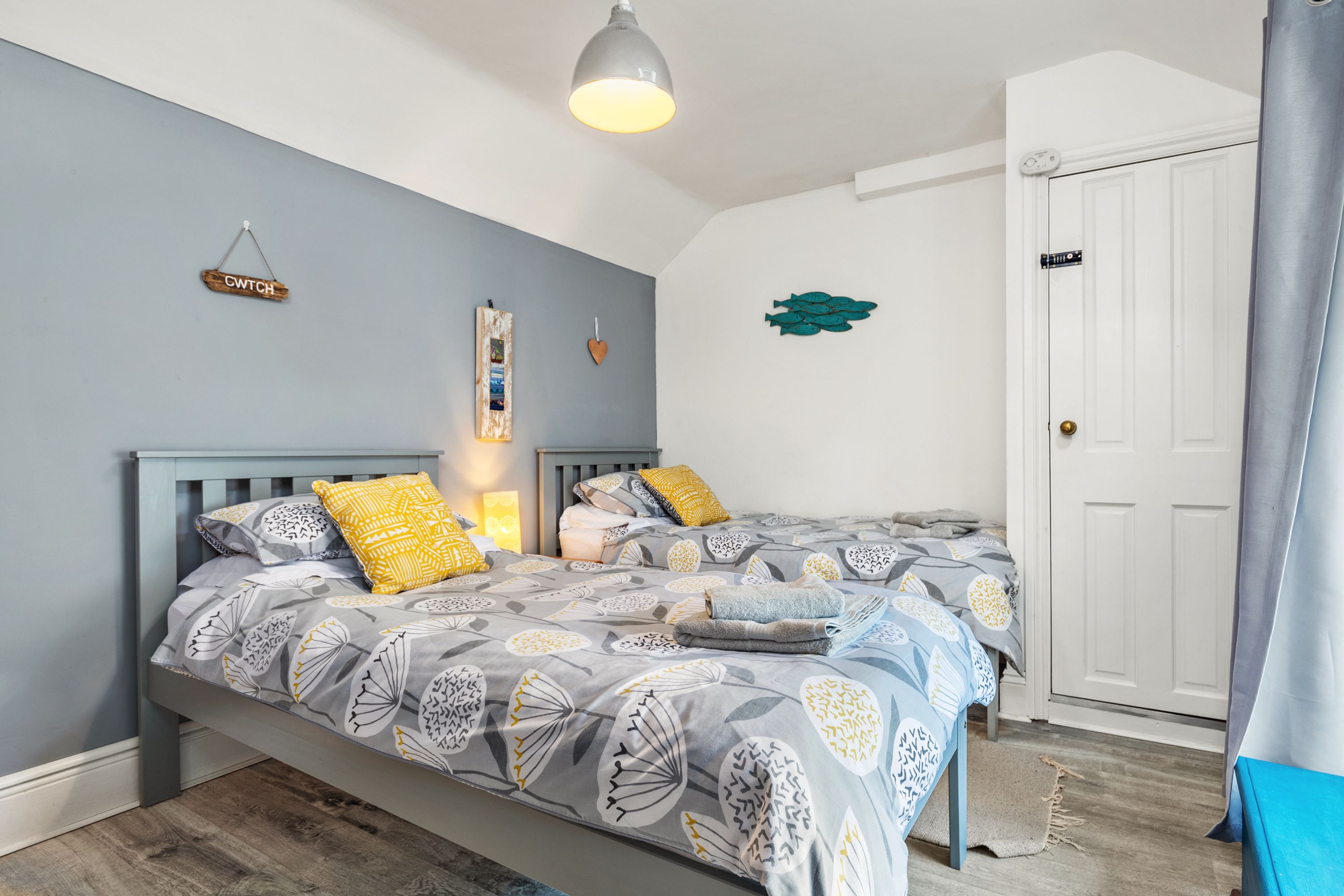 A bedroom with two beds, a grey and white patterned bedspread, a hanging light fixture, and a white door with a lock.