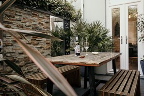 Una mesa de madera rústica con una botella y vasos, rodeada de macetas y un muro de ladrillo.