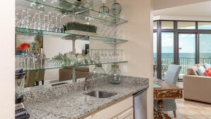 Una cocina con encimeras de granito, pileta de acero inoxidable y estantes de vidrio que exhiben copas de vino y objetos decorativos.