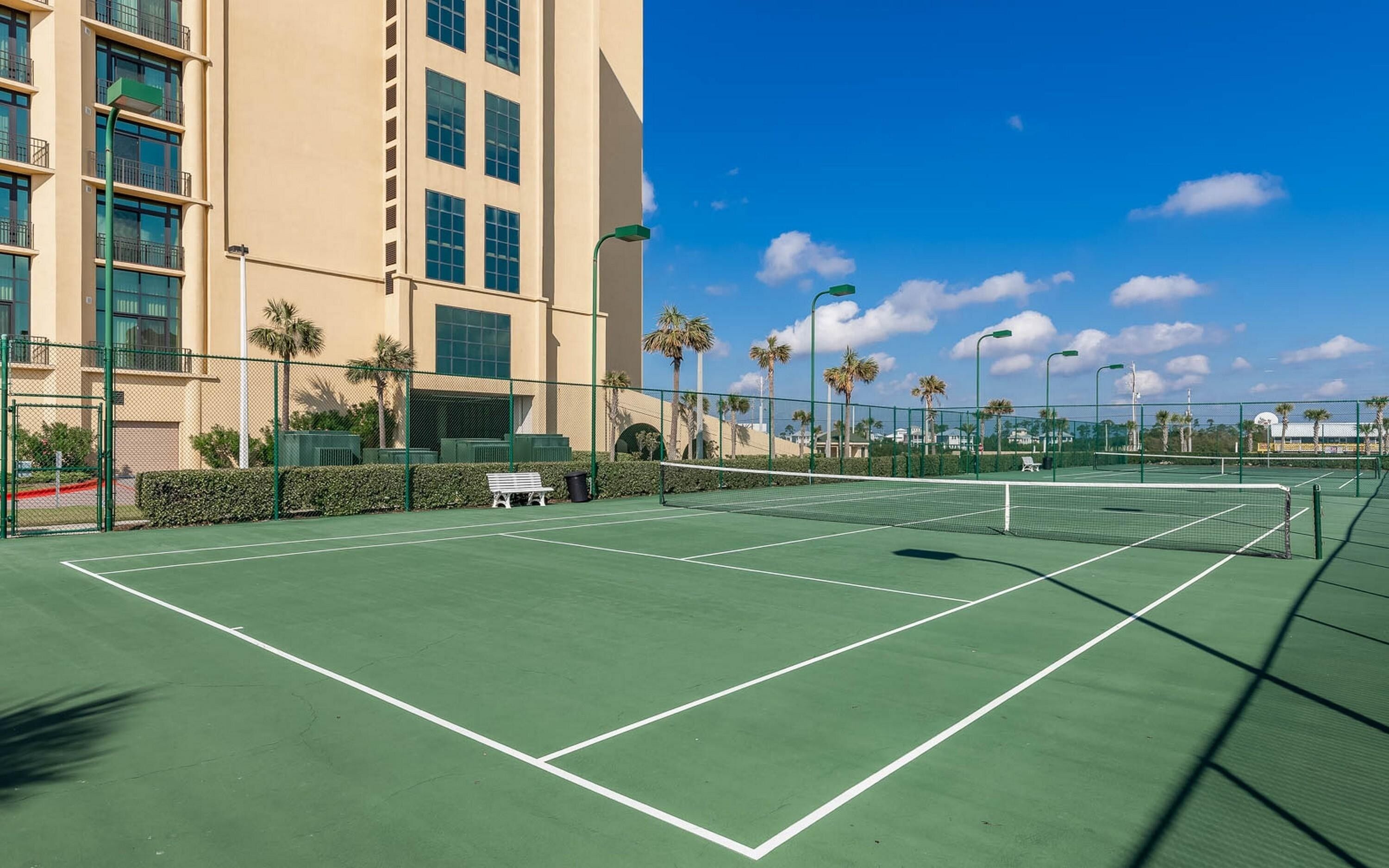 Una cancha de tenis al aire libre con una red, rodeada por una valla, y un edificio con varias ventanas al fondo.