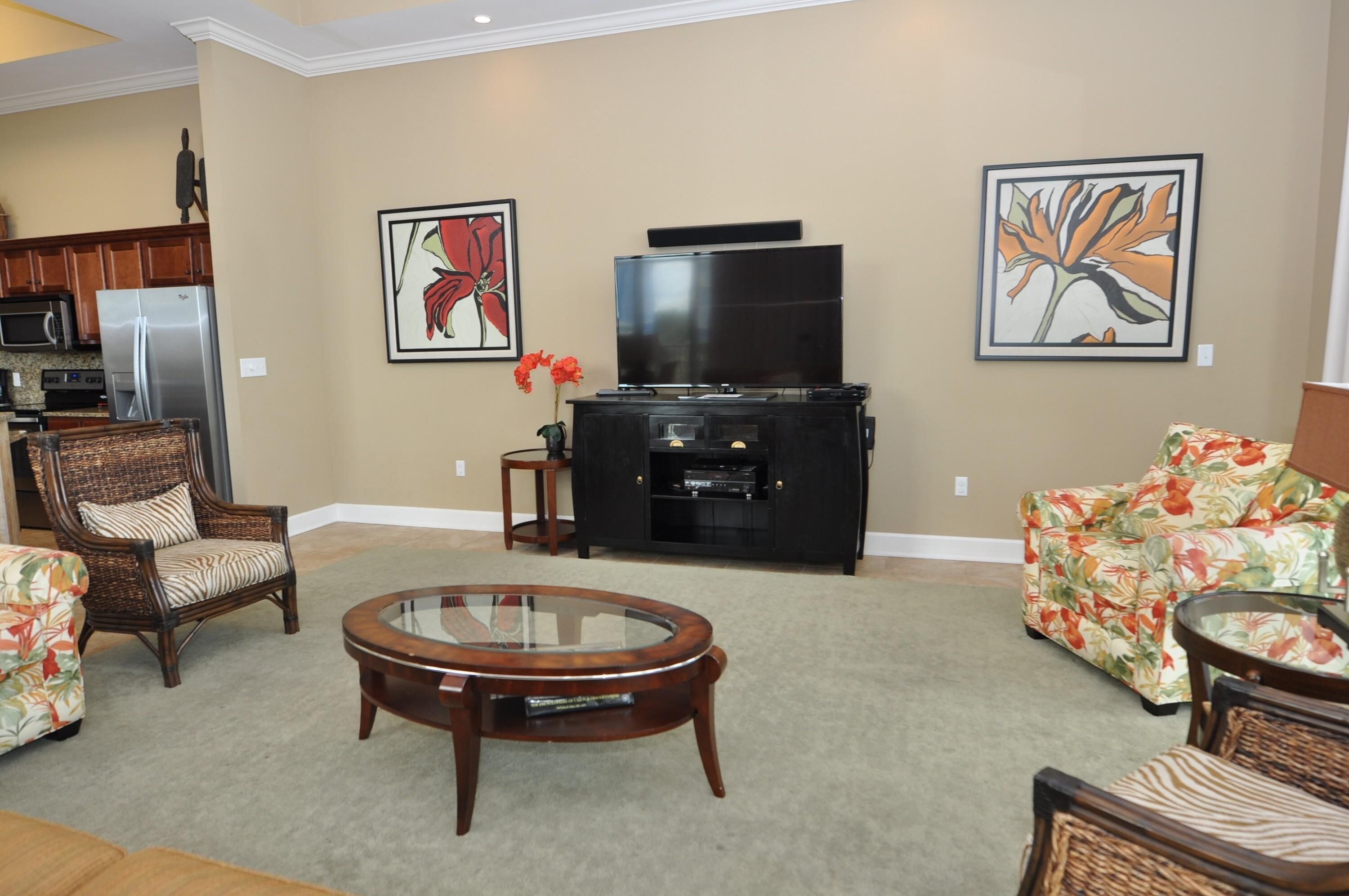 A living room with a flat-screen TV, two framed floral paintings, a wicker chair, a floral sofa, and a glass coffee table.