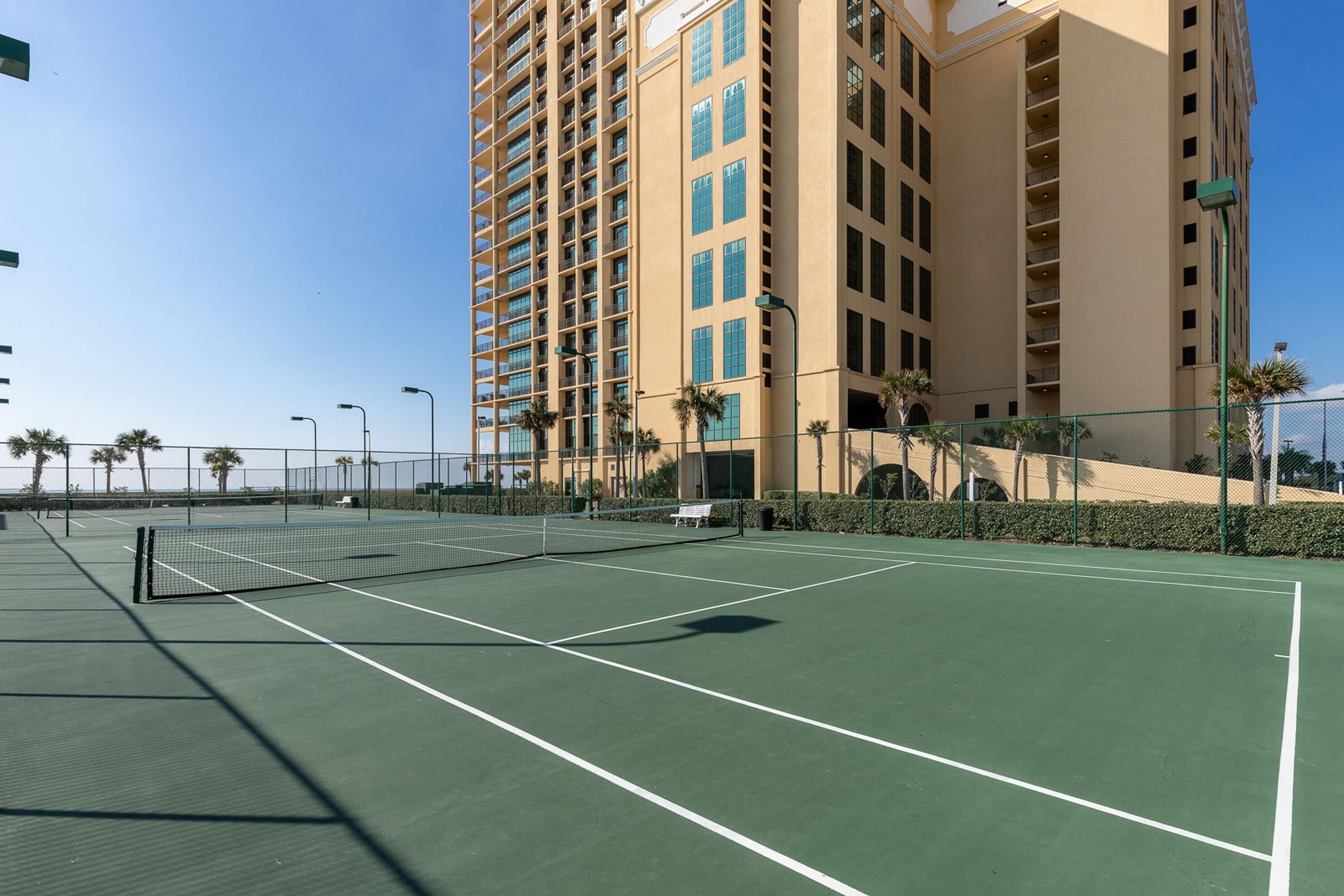An outdoor tennis court with a net, surrounded by a fence and palm trees, with a tall building in the background.