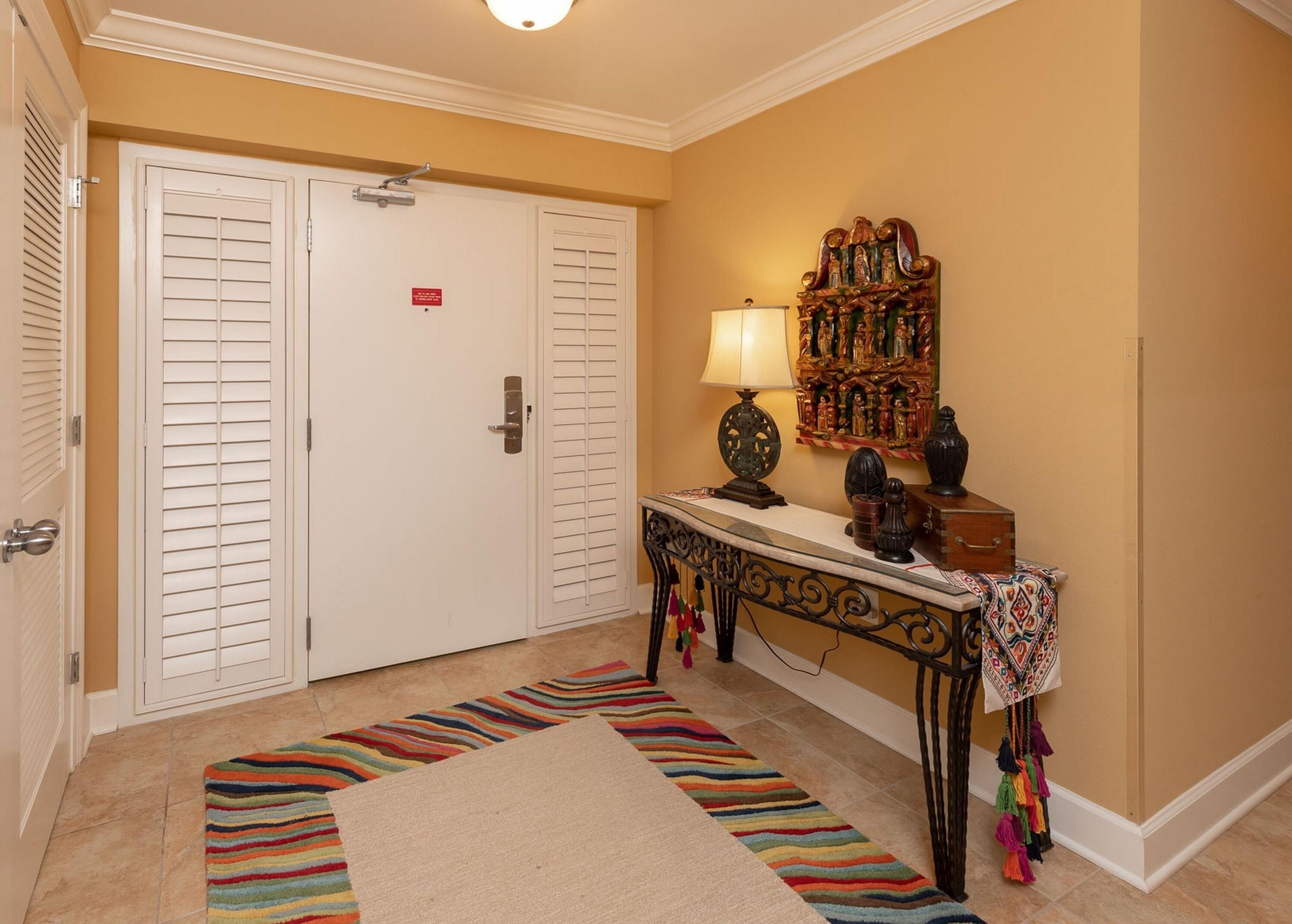 Un vestíbulo de hotel con una puerta cerrada, una consola con una lámpara, un espejo decorativo y una alfombra a rayas de colores.
