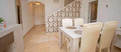 Un pasillo espacioso con suelo de mármol, una zona de comedor con una mesa y sillas blancas, y una escalera con papel tapiz decorado.