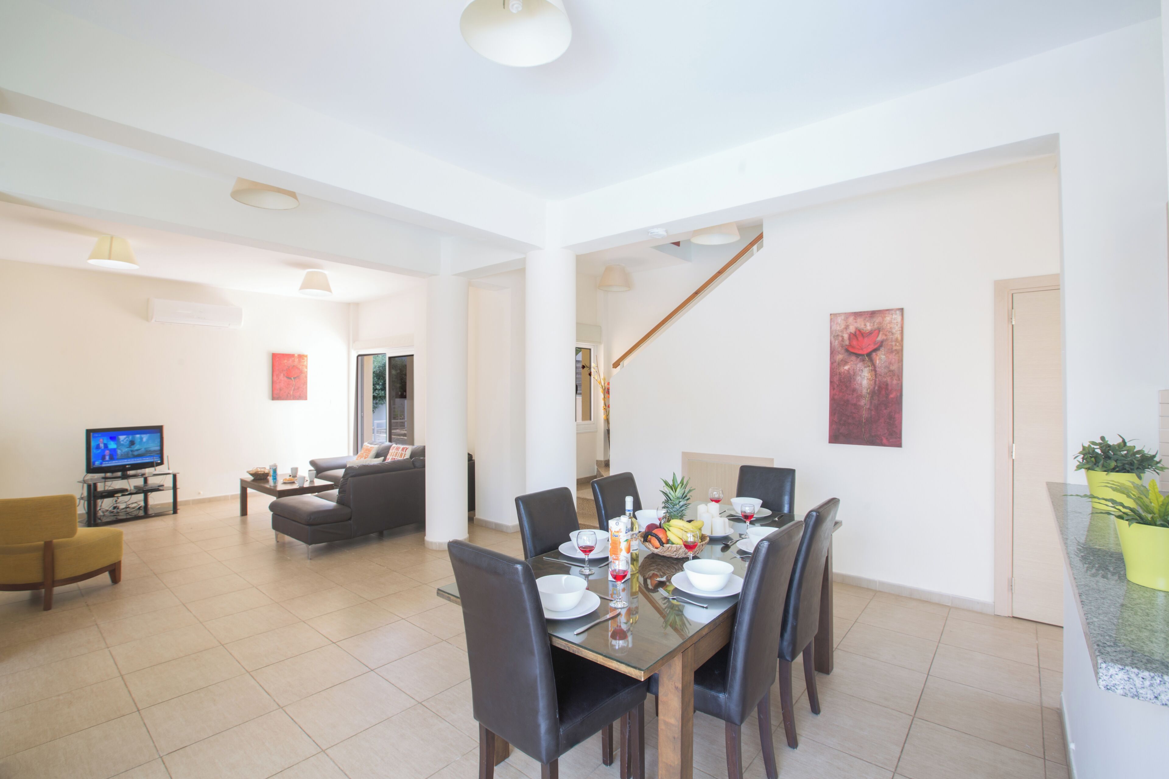 A spacious living area with a dining table set for six, a sofa, a television, and a staircase leading to another level.