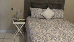 Un dormitorio con cabecera capitonada, ropa de cama estampada, una mesita de noche con lámpara y un reloj de pared.