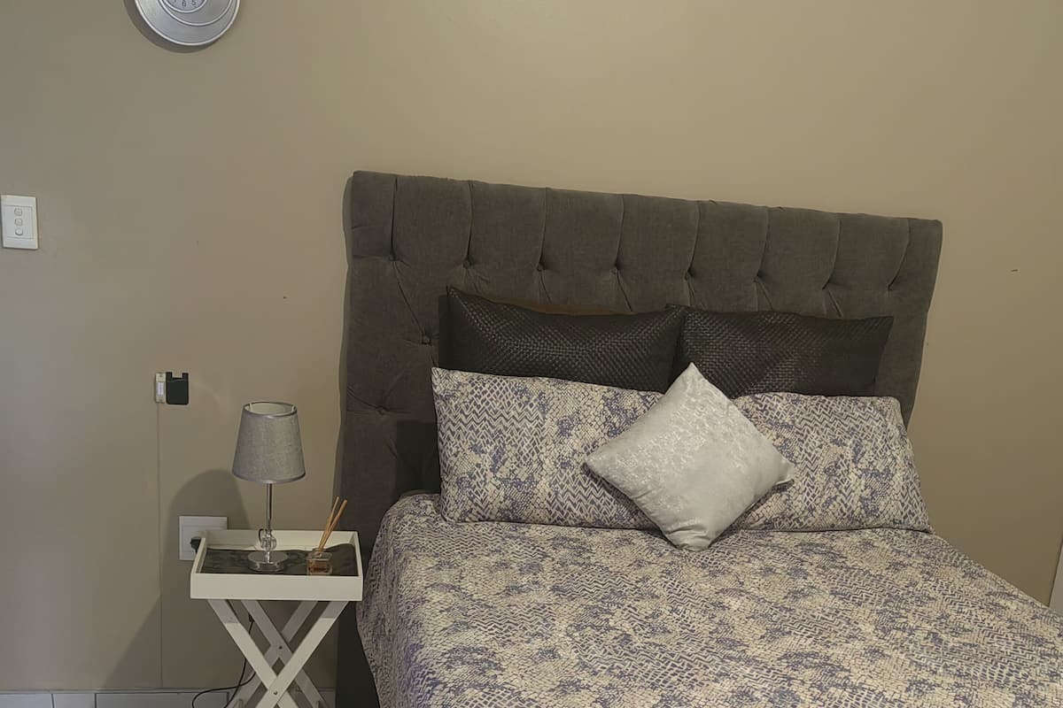 A bedroom with a tufted headboard, patterned bedding, a bedside table with a lamp, and a wall clock.