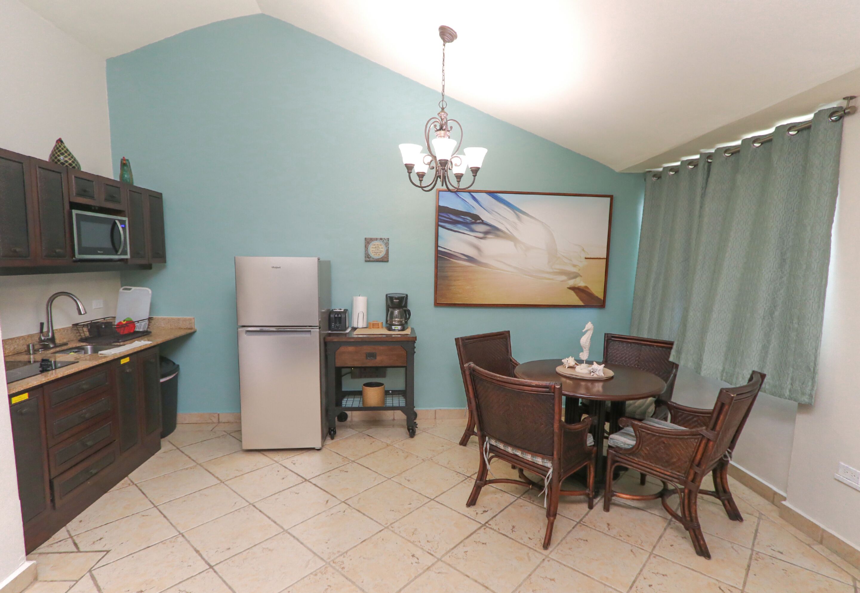 A compact kitchen with a refrigerator, microwave, and coffee maker. There's a dining table with chairs and a chandelier.