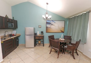 Uma cozinha compacta com geladeira, micro-ondas e cafeteira. Há uma mesa de jantar com cadeiras e um lustre.