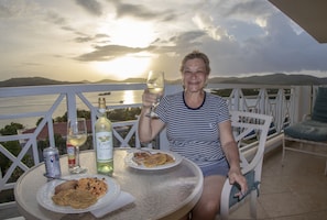 A person seated at a table with a plate of food, a glass of wine, and a bottle of Cuvée wine. The table is on an outdoor balcony with a view of a body of water and mountains in the background. - Costa Bonita Private Villa 604 (Culebra)