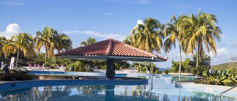 A pool area with a tiled roof structure, palm trees, and lounge chairs.