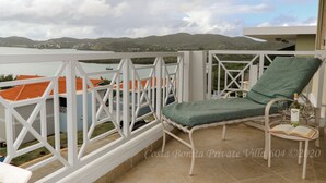Ein Balkon mit weißer Reling, einem grünen Sitzkissen und einem Tisch mit Buch und Weingläsern.