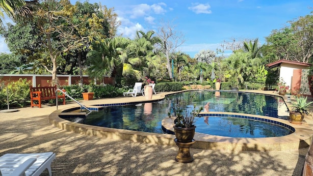 A pool area with a curved design, surrounded by lush greenery and palm trees. There are lounge chairs and a small building in the background. - Schönes Bungalow mit Gemeinschaftichem Außenpool und 2km vom Sandstrand Enfernt (Cha-am)