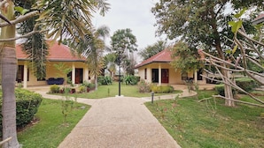 A pathway leading to two identical buildings with red-tiled roofs, surrounded by lush greenery and palm trees. - Schönes Bungalow mit Gemeinschaftichem Außenpool und 2km vom Sandstrand Enfernt (Cha-am)