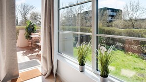 A room with a large window, a wooden floor, a potted plant, and a view of a garden and buildings outside. - Charm 1 - Chic 2-bdr Apt With Shared Garden (Copenhagen)