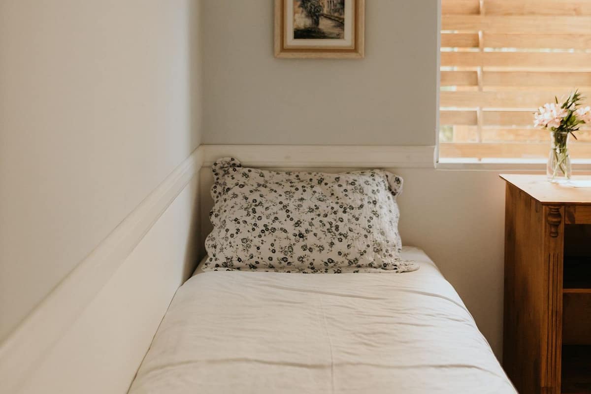 A small, neatly made bed with a floral-patterned bedspread and a matching pillow, a framed picture on the wall, and a window with a wooden blind.