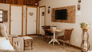A cozy room with wooden walls, a white ceiling, a flat-screen TV mounted on a wooden panel, a dining table with chairs, and a small side table with a plant. - Paty's Garden (Florianópolis)