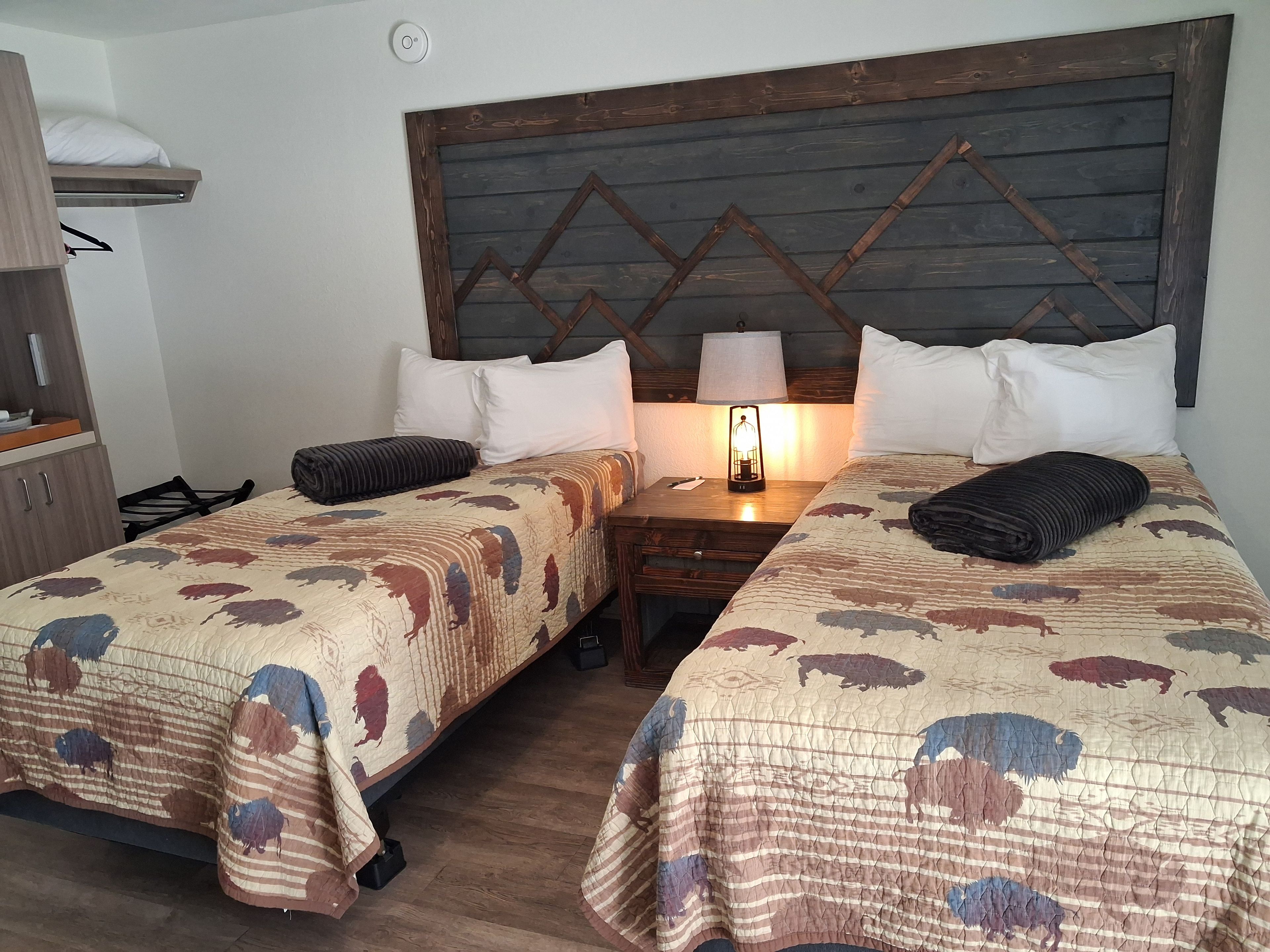 Habitación de hotel con dos camas, cabecero de madera, mesita de noche con lámpara y una pequeña mesa.