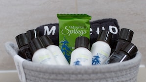 Una cesta de mimbre gris con varios artículos de tocador personal, incluyendo una botella de loción Mountain Springs verde, sobre una superficie de azulejos.