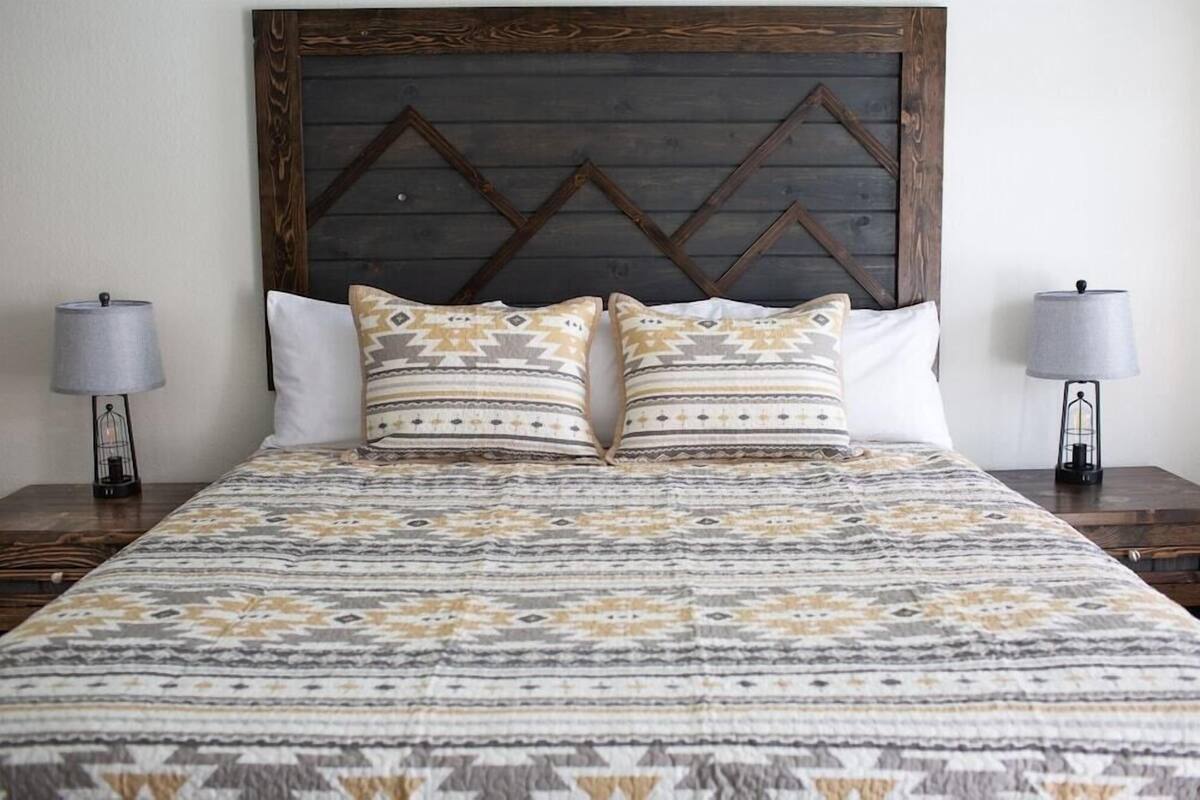 Una cama con una colcha estampada, dos almohadas con diseños geométricos y un cabecero de madera con un patrón de espiga.