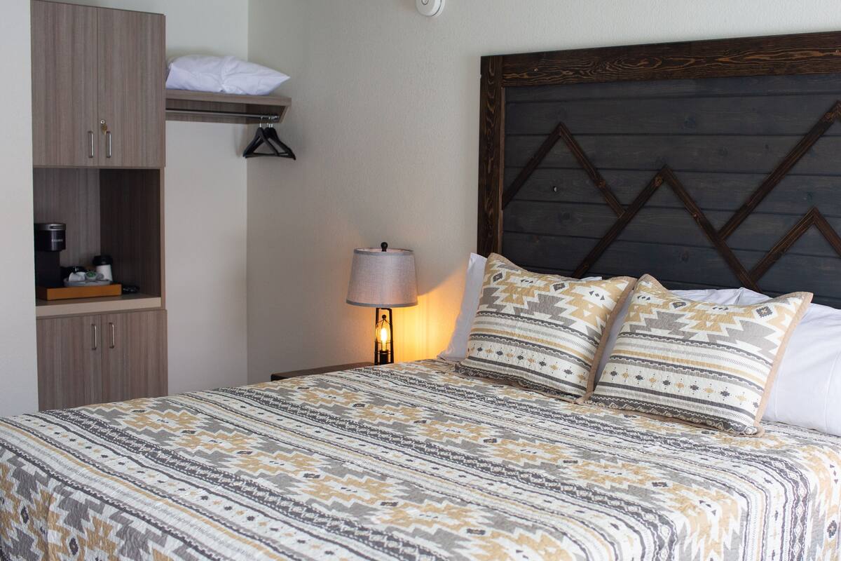 Una cama bien hecha con ropa de cama estampada, una mesita de noche con una lámpara, un cabecero de madera y un armario con repisas colgantes.