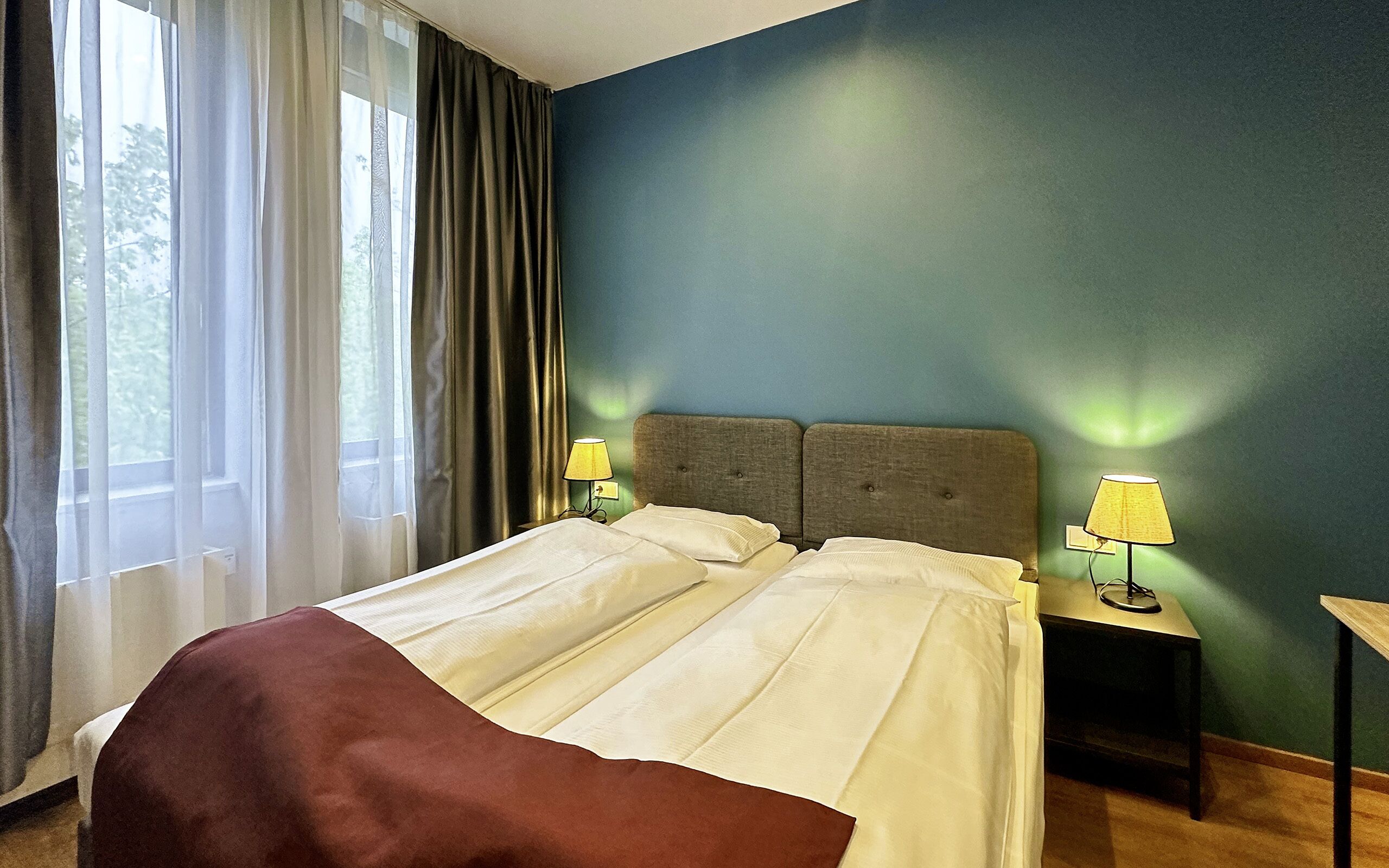 A hotel room with a bed, two bedside lamps, a window with curtains, and a green wall.