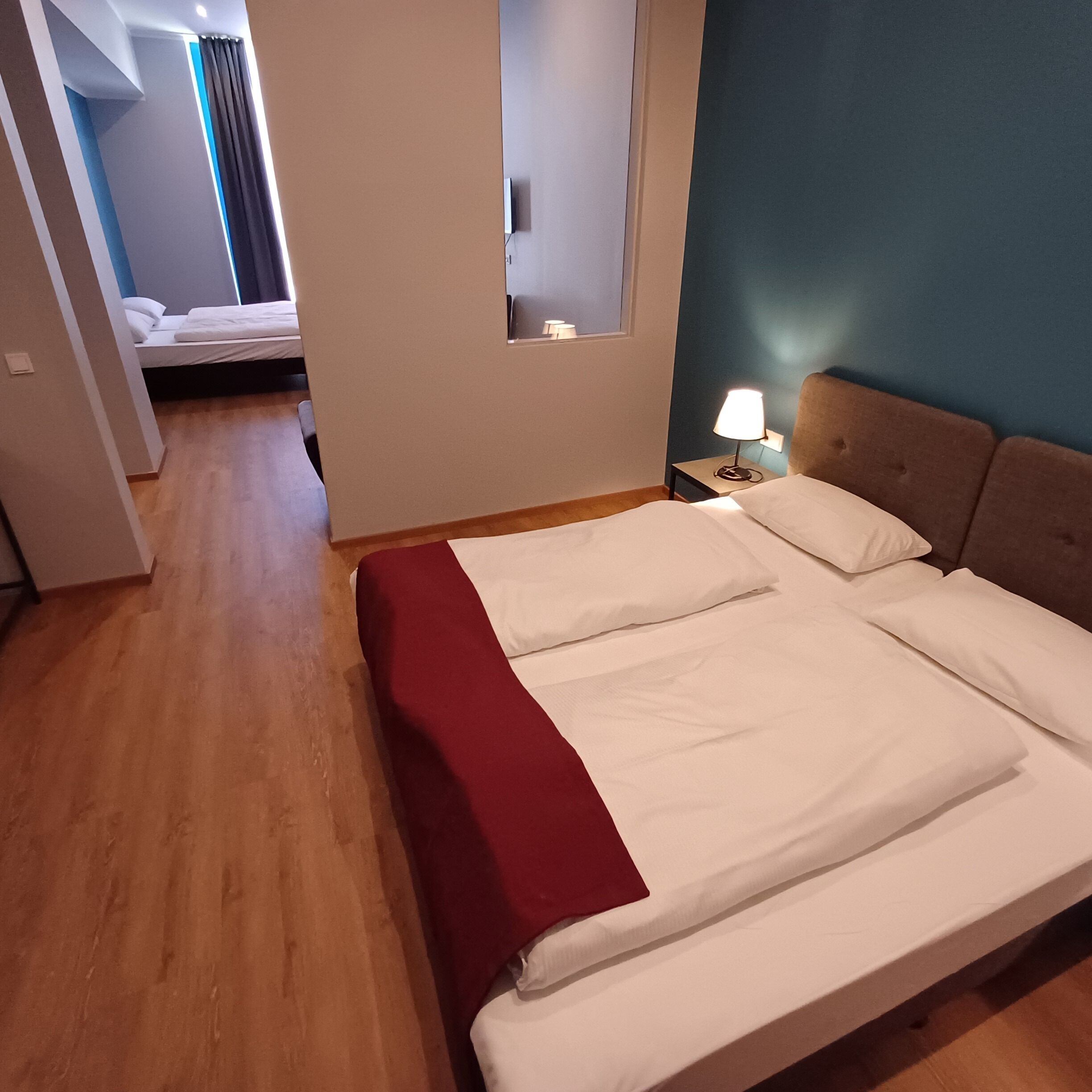 A hotel room with a bed, a nightstand with a lamp, a wooden floor, and a view of another room with a bed.