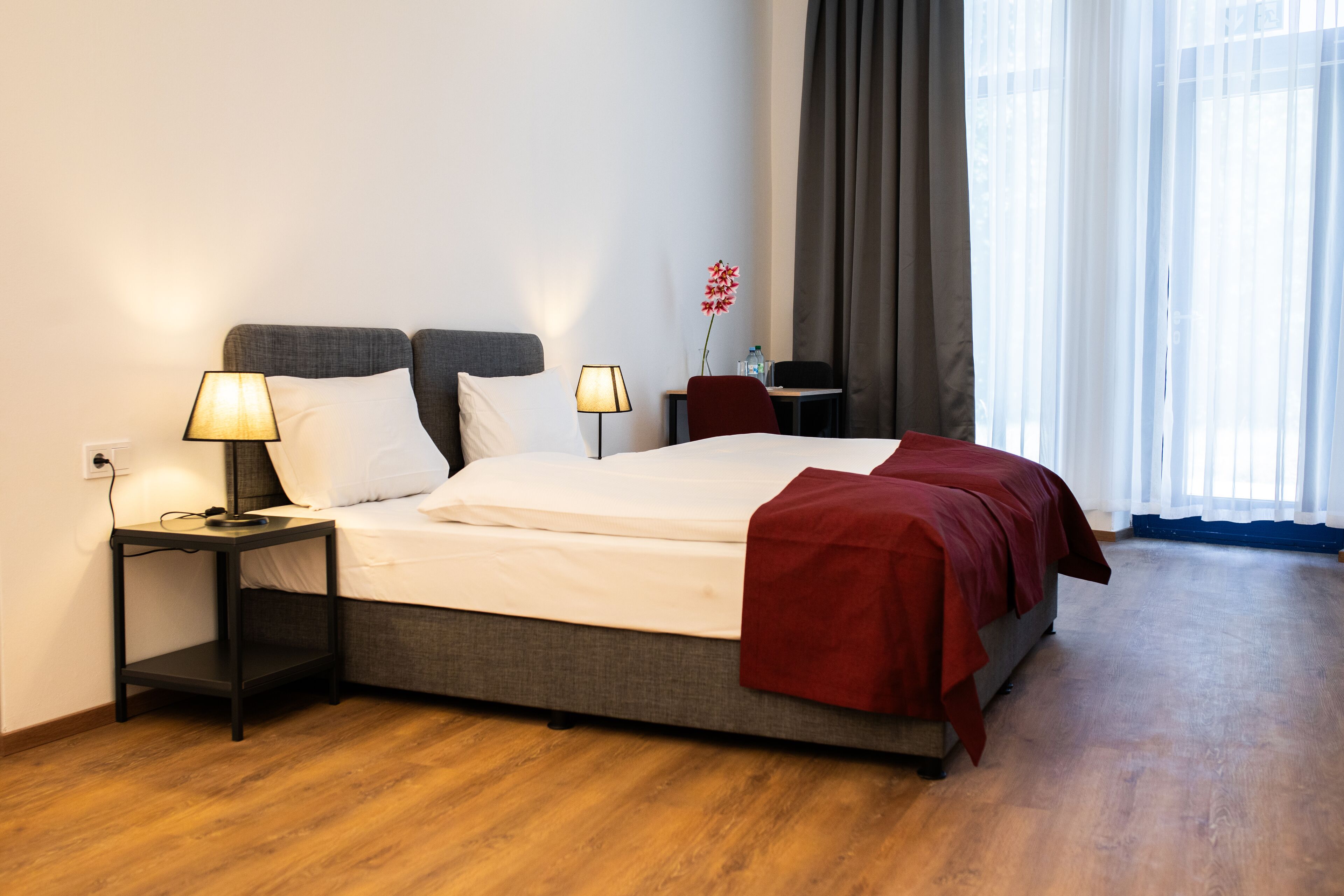 Een keurig opgemaakt bed met een rode deken, twee bedlampen en een klein tafeltje met een stoel.