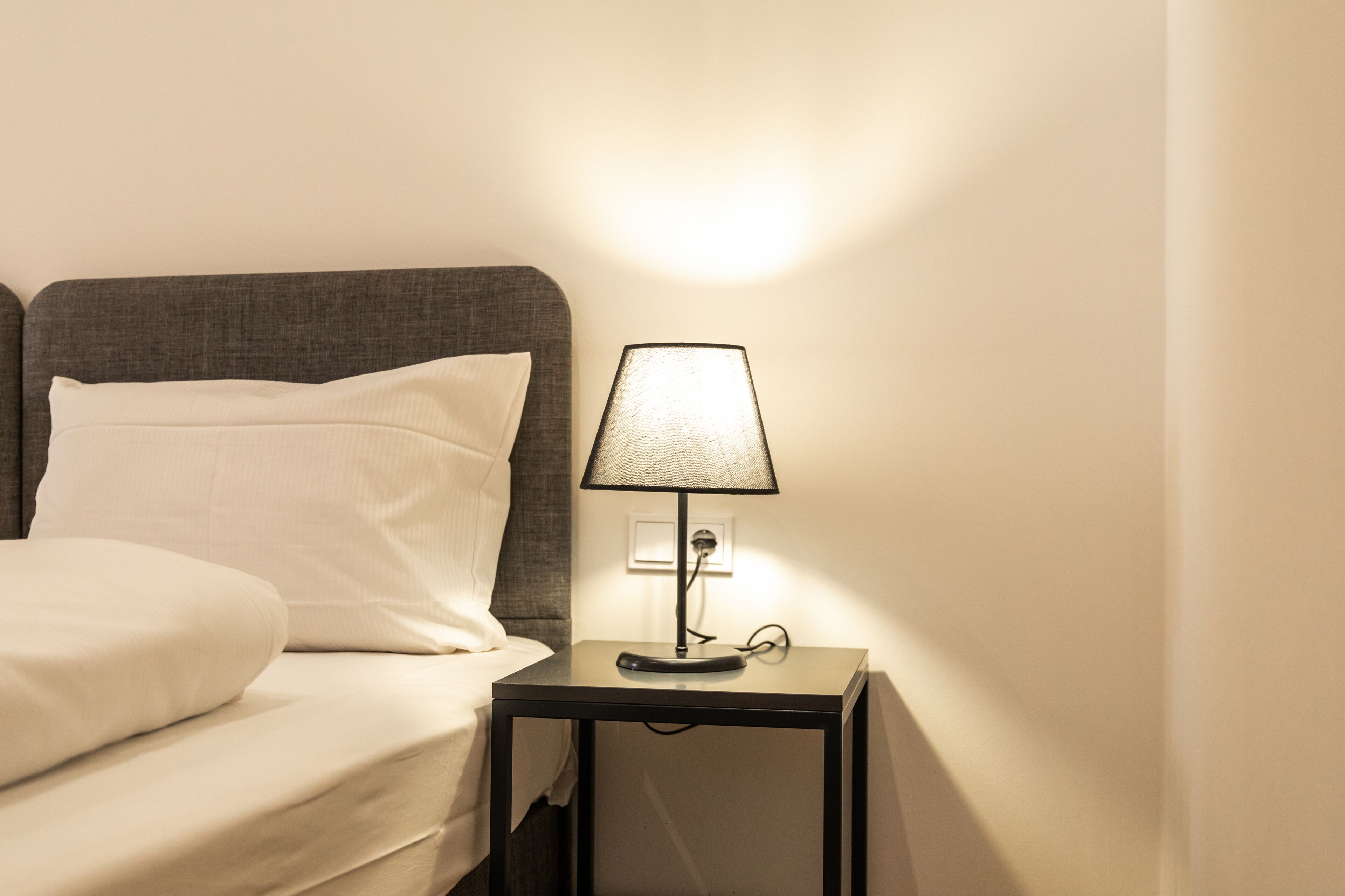 A neatly made bed with white linens and a pillow, a small bedside table with a lamp, and a power outlet on the wall.