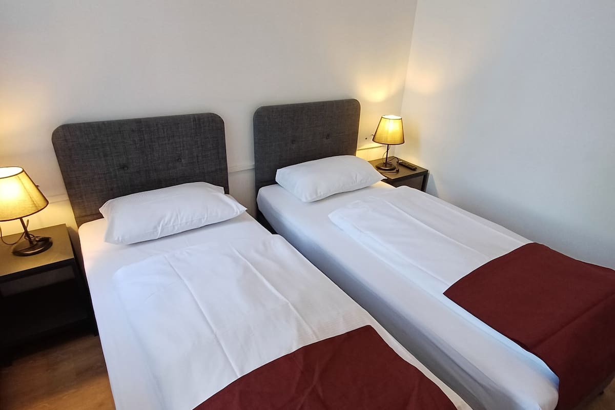Quarto de hotel com duas camas, cada uma com cabeceira cinza e roupa de cama branca. Há mesas de cabeceira com luminárias e um controle remoto em uma delas.