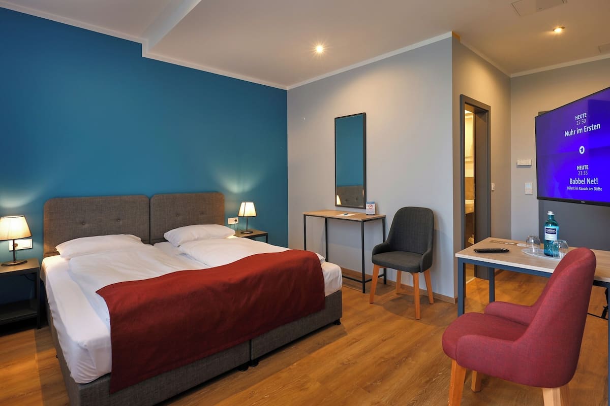 Quarto de hotel com uma cama, uma escrivaninha, uma cadeira e uma televisão mostrando um programa.