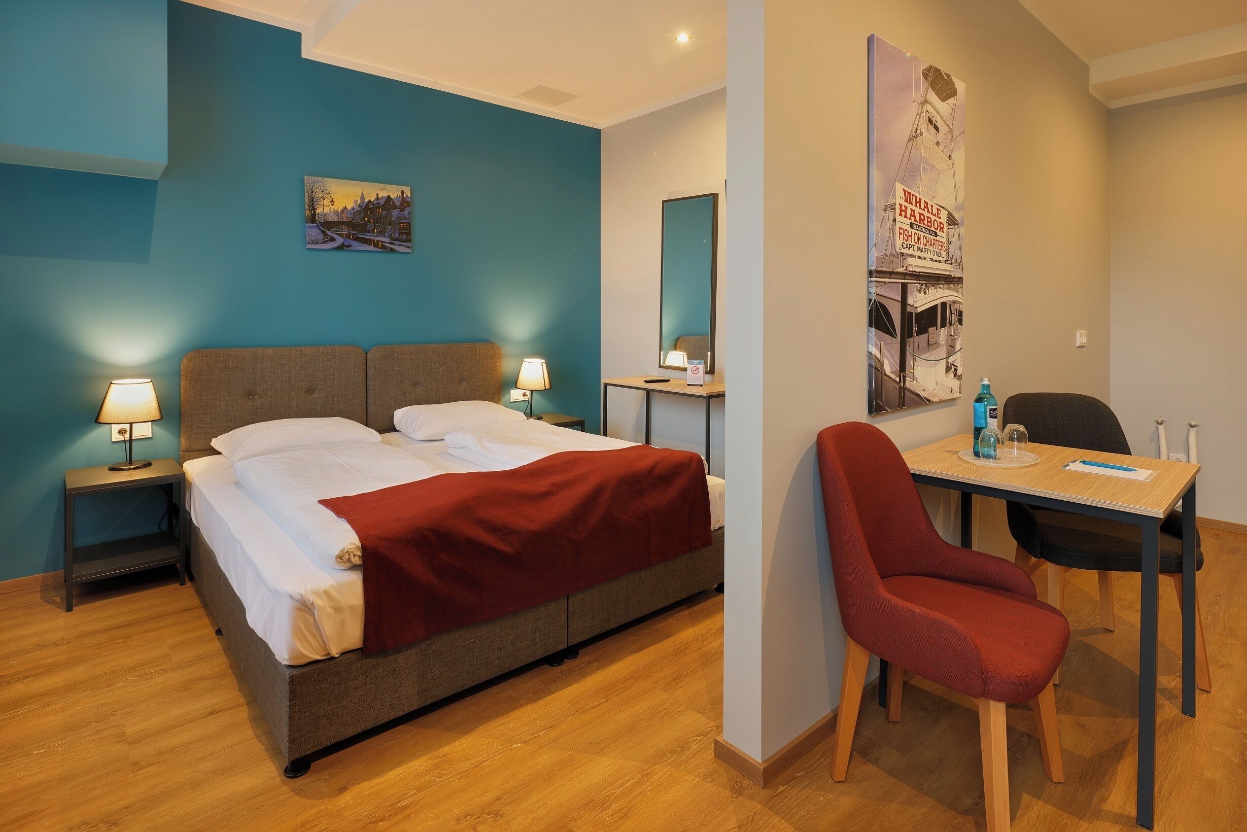 A hotel room with a bed, bedside tables, a desk with a chair, a mirror, and a wall with a poster.