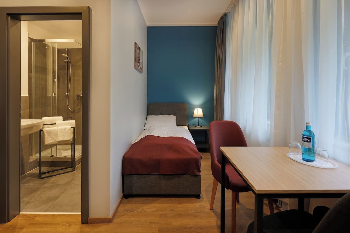 Quarto de hotel com cama, escrivaninha e cadeira, mesa com garrafa, mesa de cabeceira com abajur e banheiro com chuveiro e pia.