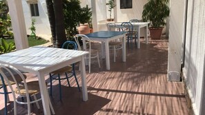 Un patio exterior con mesas y sillas blancas, rodeado de plantas en macetas y un edificio blanco al fondo.