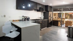 A modern kitchen with a central island, dark cabinetry, and a view into a living area with a pool. - Modern Villa Steps to Nightlife (Jacó)