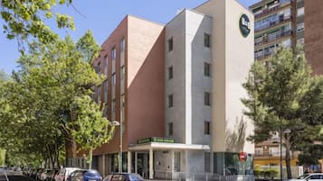 Un hotel de varios pisos con una imponente torre de reloj, un estacionamiento con varios coches y una calle arbolada.